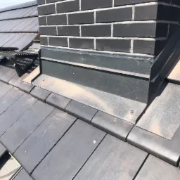 Szare dachówki ceramiczne wokół komina z czarnej cegły, zabezpieczone obróbką blacharską, widoczna ława kominiarska z metalową kratką.