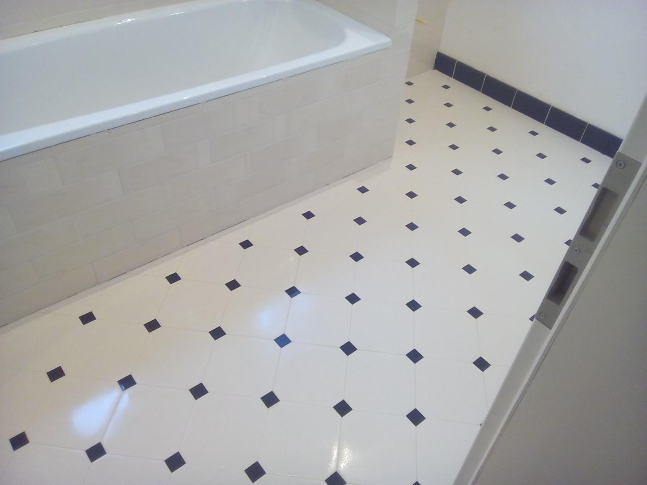 Wykończona łazienka z białą wanną obłożoną kafelkami imitującymi cegłę i białą podłogą z regularnym wzorem czarnych kwadratów, widoczny fragment drzwi.