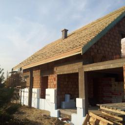 MTBud - Wznoszony dom z czerwonej cegły z widocznym zadaszeniem i kominem, ocieplony styropianem przy fundamencie, w tle drzewa i rusztowanie.