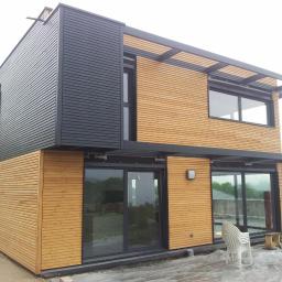 MTBud - Nowoczesny dom z elementami drewnianymi i czarnymi panelami, duże przesuwne okna, widoczne prace wykończeniowe na zewnątrz, w tle zielony krajobraz.