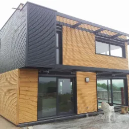 Nowoczesny dom z elementami drewnianymi i czarnymi panelami, duże przesuwne okna, widoczne prace wykończeniowe na zewnątrz, w tle zielony krajobraz.