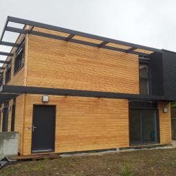 MTBud - Nowoczesny dwukondygnacyjny dom z elewacją z jasnego drewna, czarnymi ramami okiennymi i metalową konstrukcją nadającą charakterystyczny wygląd, widoczny z zewnątrz w pochmurny dzień.