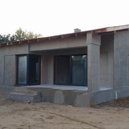 MTBud - Surowy, jednokondygnacyjny dom w trakcie budowy, z widocznymi oknami i drzwiami balkonowymi, otoczony piaszczystym terenem i drzewami.