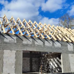 Budowa domu murowanego w stanie surowym otwartym: ściany z bloczków, drewniana konstrukcja dachu widoczna na tle błękitnego nieba z chmurami.