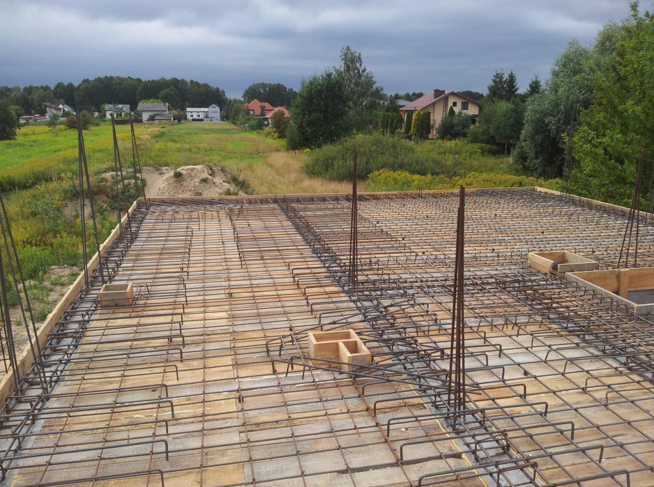 Zbrojenie płyty fundamentowej na drewnianym szalunku, widoczne wystające pręty i drewniane formy, w tle krajobraz z zabudowaniami i pochmurne niebo.