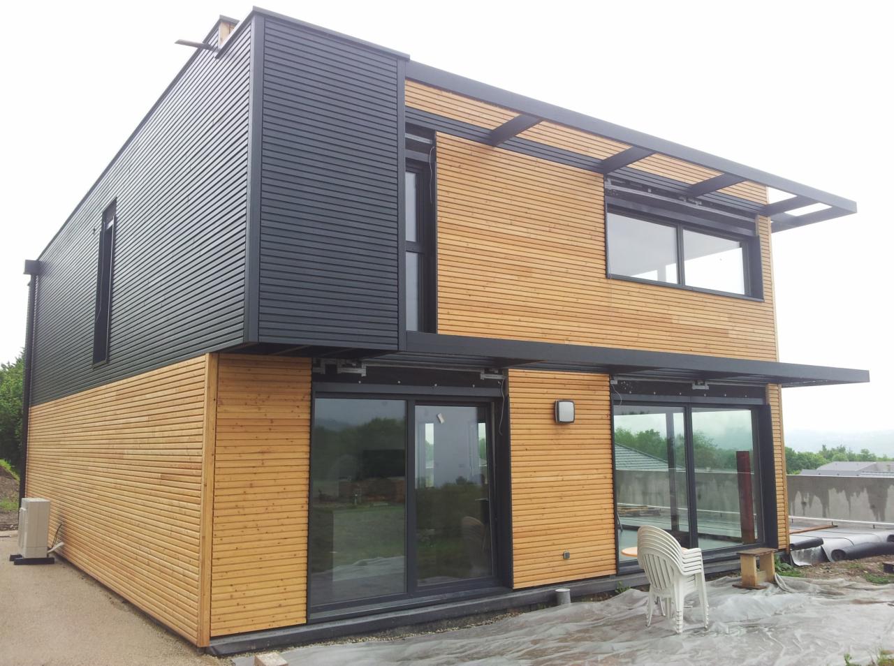 Nowoczesny dom z elementami drewnianymi i czarnymi panelami, duże przesuwne okna, widoczne prace wykończeniowe na zewnątrz, w tle zielony krajobraz.