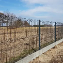 Długie, szare ogrodzenie panelowe z podmurówką betonową, widok z boku, pole w tle.