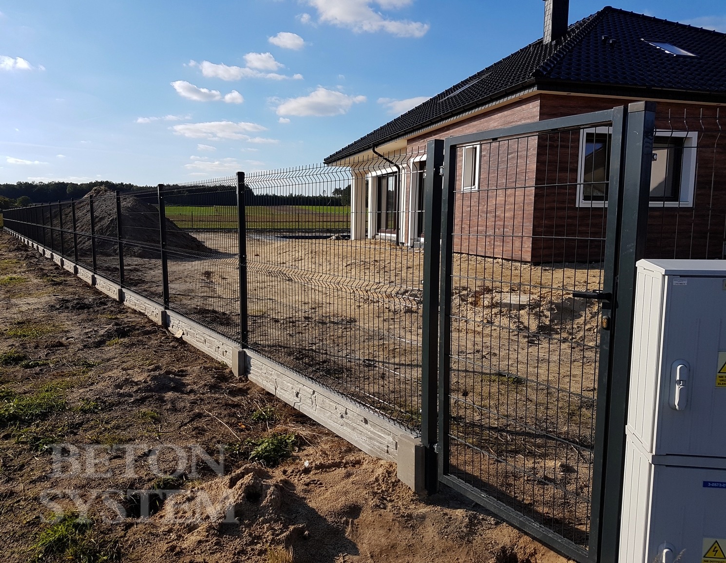 Nowoczesne ogrodzenie panelowe z podmurówką betonową i furtką w Ujeździe, widoczne na tle nowo wybudowanego domu z brązową elewacją i stertą ziemi wzdłuż ogrodzenia.