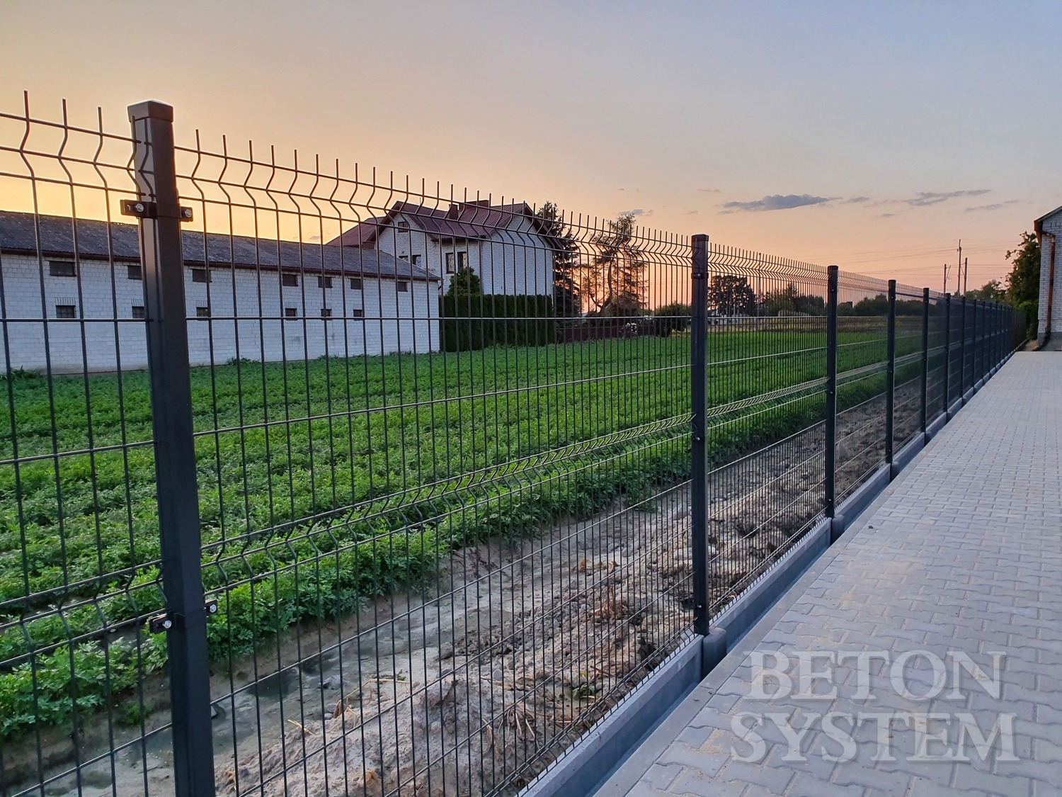 Długie, proste, ciemne ogrodzenie z siatki panelowej, widok z boku, w tle budynki gospodarcze i dom jednorodzinny o zachodzie słońca.