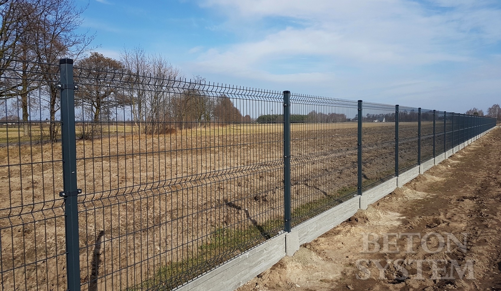 Długie, szare ogrodzenie panelowe z podmurówką betonową, widok z boku, pole w tle.