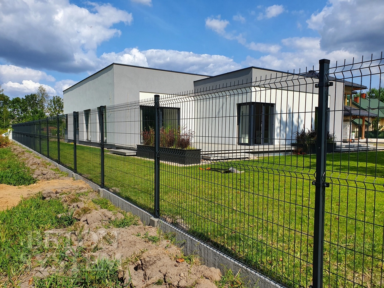 Nowoczesne, antracytowe ogrodzenie z paneli kratowych na betonowej podmurówce, oddzielające posesję z minimalistycznym domem i zadbanym trawnikiem od niezagospodarowanego terenu.