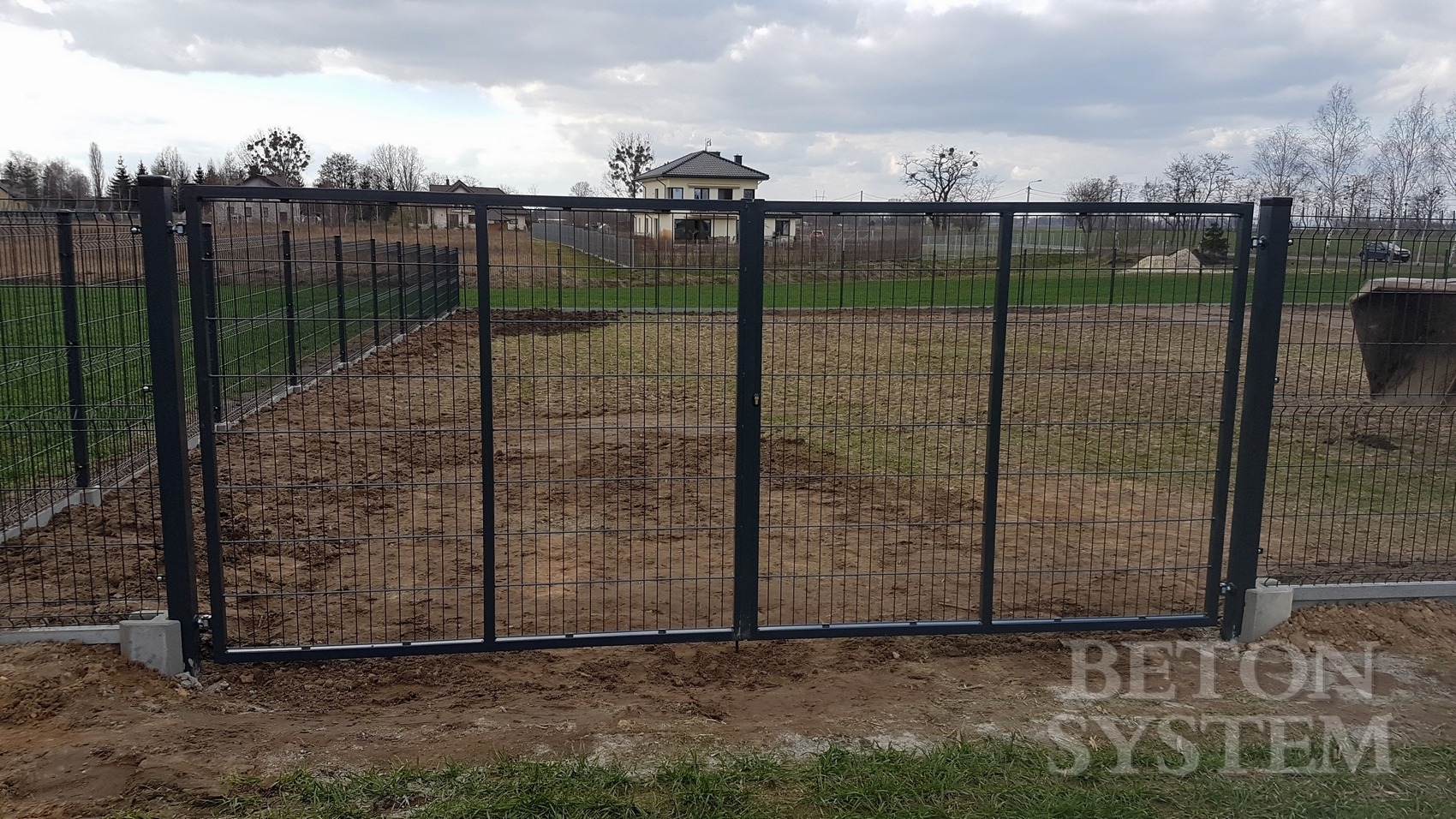 Nowoczesna brama dwuskrzydłowa z siatki z metalowymi słupkami, osadzona na betonowej podmurówce, z widocznym domem w tle i pochmurnym niebem.
