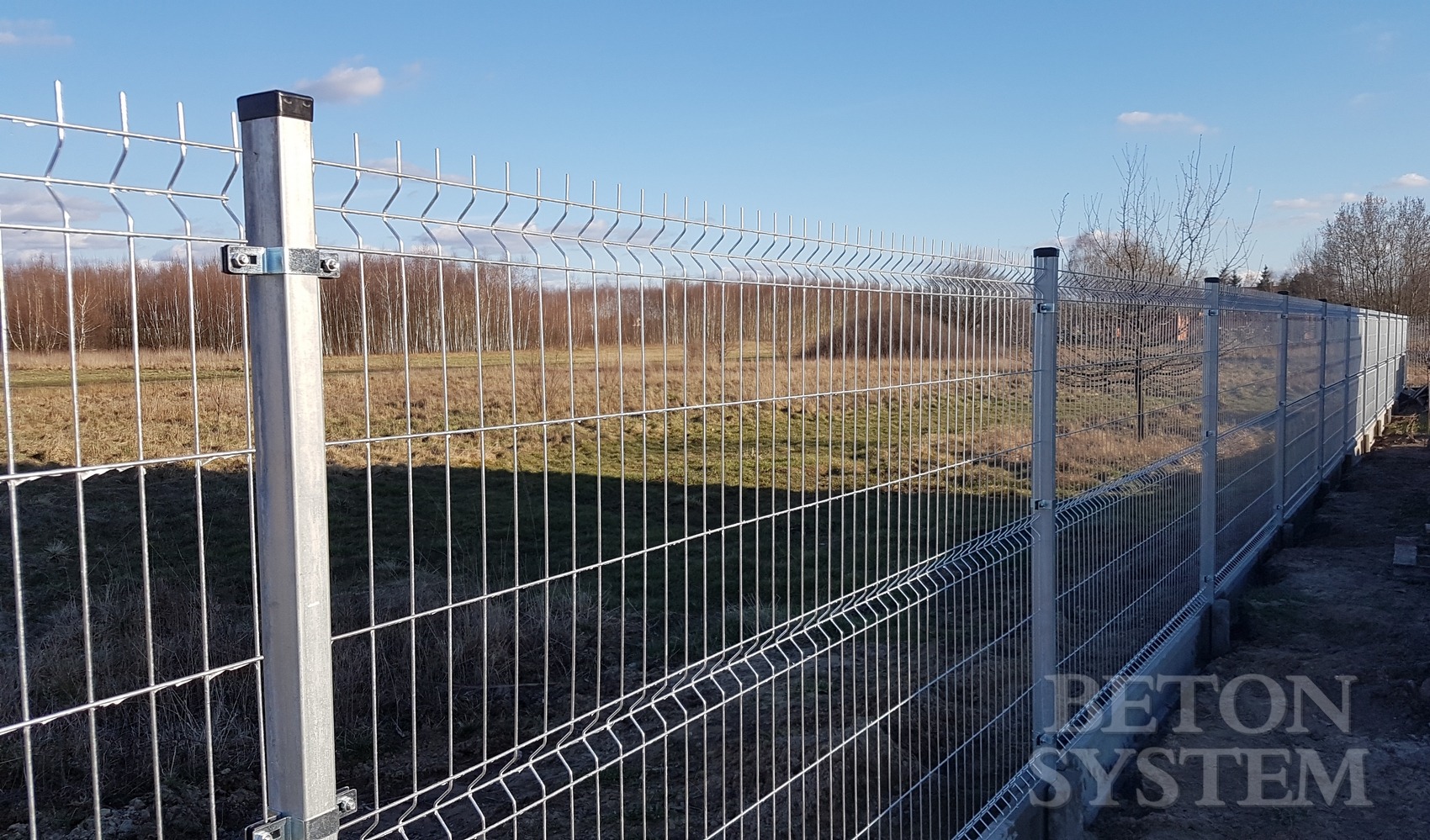 Długi odcinek nowoczesnego ogrodzenia panelowego z metalowymi słupkami i złączkami, widoczne pole i drzewa w tle, jasne światło słoneczne.