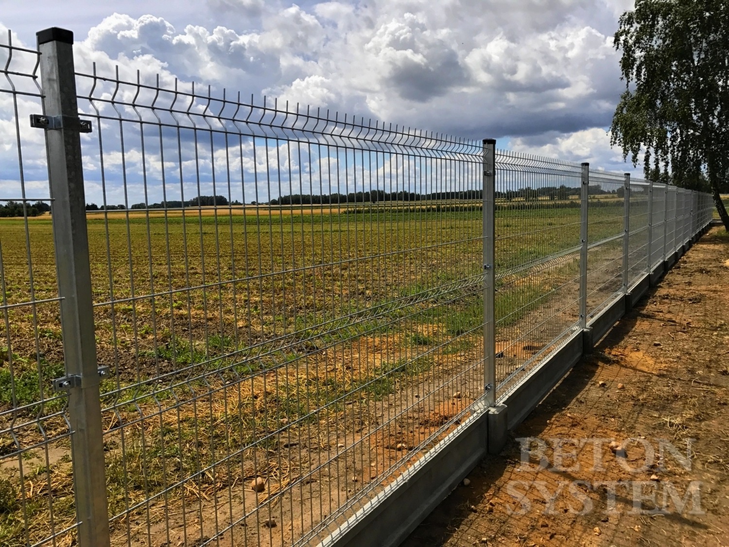 Długie, proste ogrodzenie z siatki z metalowymi słupkami i betonową podmurówką, rozciągające się wzdłuż pola uprawnego pod pochmurnym niebem.