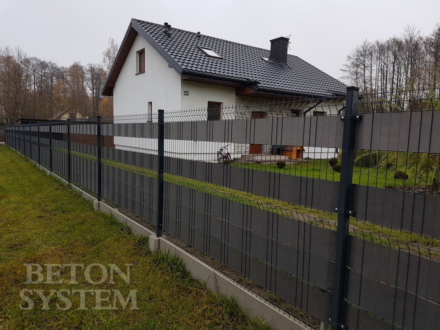 Długie, nowoczesne ogrodzenie z paneli w kolorze grafitowym, z siatką na górze i betonową podmurówką, biegnące wzdłuż trawnika przed domem jednorodzinnym z czarnym dachem.