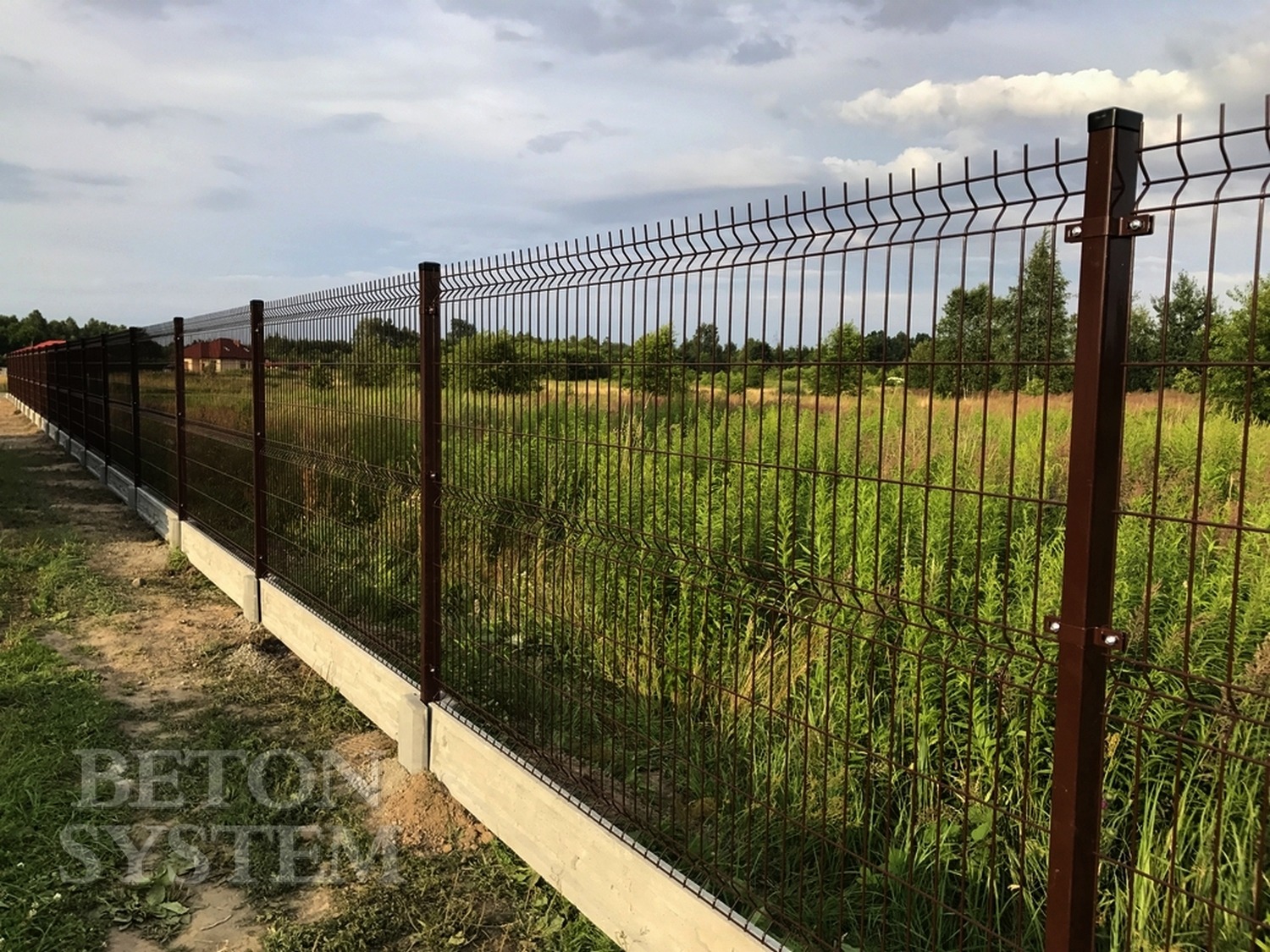 Długie, brązowe ogrodzenie z paneli z podmurówką betonową, widoczne z boku na tle łąki i drzew.