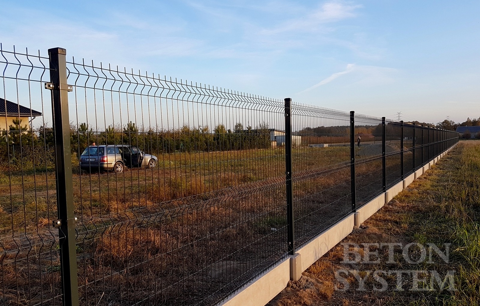 Długi, czarny system ogrodzeniowy z paneli na betonowej podmurówce, biegnący wzdłuż pola z zaparkowanym samochodem osobowym z otwartymi drzwiami w tle, widok z perspektywy ziemi.