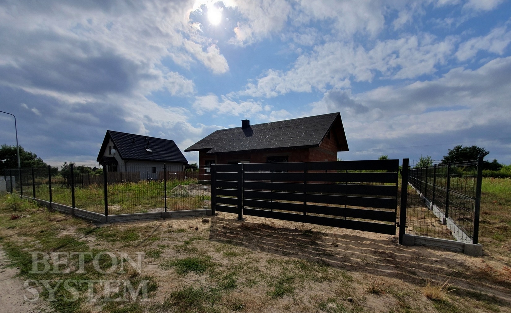 Nowoczesne ogrodzenie posesji z czarnych, poziomych paneli metalowych i siatki, z widocznymi dwoma domami w tle, pod pochmurnym niebem.
