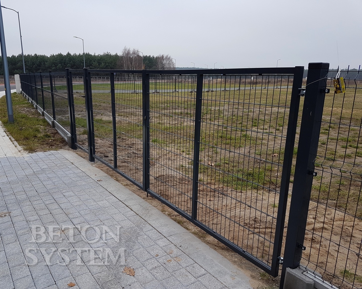 Ciemnoszare ogrodzenie z siatki z bramą, widok z bliska na kostkę brukową i trawnik, w tle las i słupy oświetleniowe.