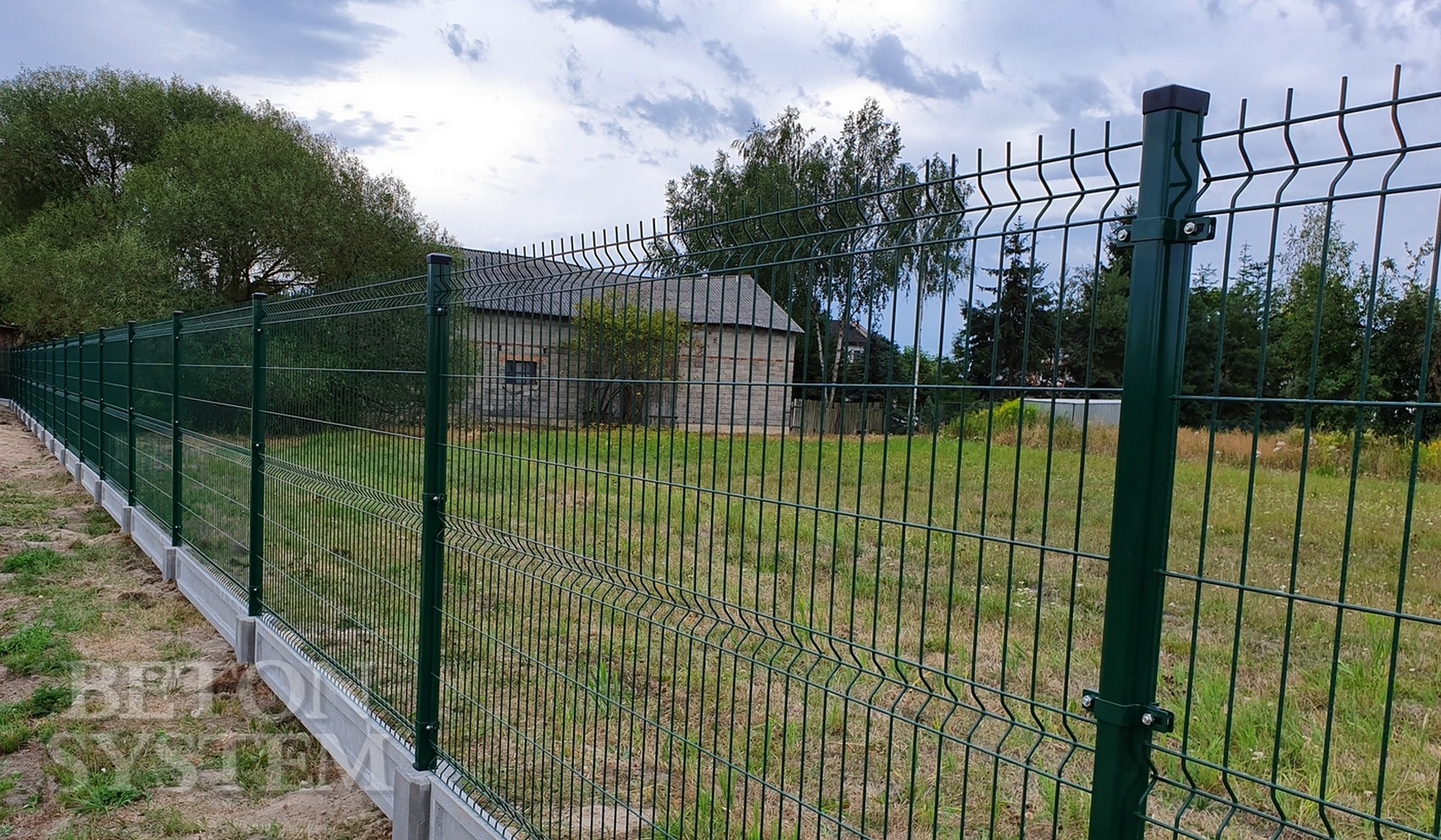 Zielone ogrodzenie panelowe na betonowej podmurówce, widok z zewnątrz posesji, w tle budynek gospodarczy.