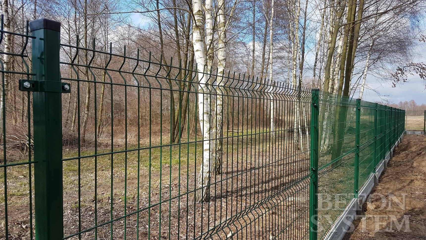 Zielone, stalowe ogrodzenie panelowe na betonowej podmurówce, widok z bliska na słupki i siatkę z zagiętymi prętami, w tle drzewa bez liści.