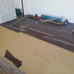 Częściowo ułożona podłoga z paneli w odcieniach brązu, widoczne narzędzia pomiarowe, paczki paneli w tle oraz fragment tapety w geometryczne wzory na ścianie.