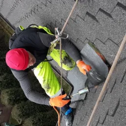 Pracownik w czerwonej czapce i jaskrawych spodniach, zabezpieczony linami, lutuje papę na dachu krytym gontem bitumicznym, używając palnika.