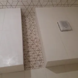 Białe płytki ścienne z geometrycznym wzorem, fragment zabudowy z wpuszczonym panelem sterowania, widoczne fugi i krawędzie płytek.