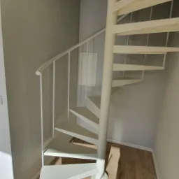 Białe metalowe schody spiralne w jasnym wnętrzu, widok z dołu, drewnopodobna podłoga, minimalistyczny design.