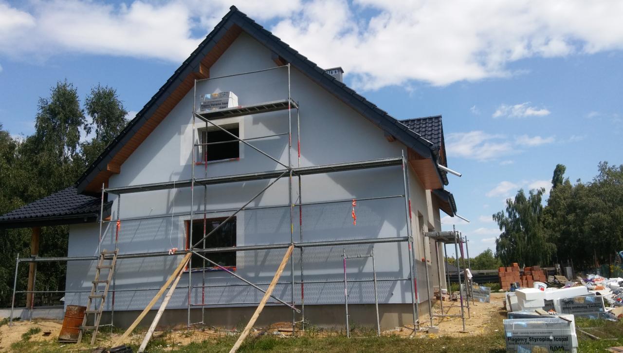 Dom jednorodzinny w trakcie ocieplania, widoczne rusztowania, siatka elewacyjna, oraz materiały izolacyjne obok budynku.