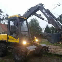 Żółto-szara koparka Mecalac MXT na nierównym terenie, z włączonymi światłami, w tle dom z czerwonym dachem i sterta drewna za ogrodzeniem.