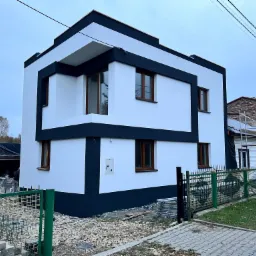 Nowoczesny, dwukondygnacyjny dom w Maszkowicach z białą elewacją obramowaną czarnymi pasami, widok z zewnątrz na tle sąsiednich budynków i zielonego ogrodzenia.