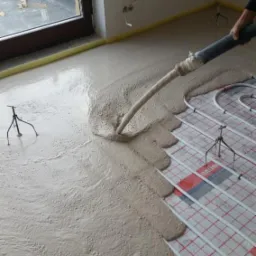 Wylewka płynna w trakcie rozprowadzania na ogrzewaniu podłogowym, widoczne rury grzewcze i podpory, żółta taśma dylatacyjna przy ścianie.