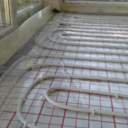 Rozłożone rury ogrzewania podłogowego na folii z siatką, widoczne okno z profilem Alumander i taśma klejąca.