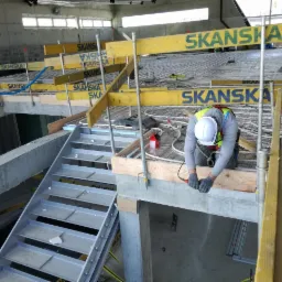 Pracownik w kasku szlifuje betonową powierzchnię, widoczne rury ogrzewania podłogowego i konstrukcja wsporcza z logo Skanska.
