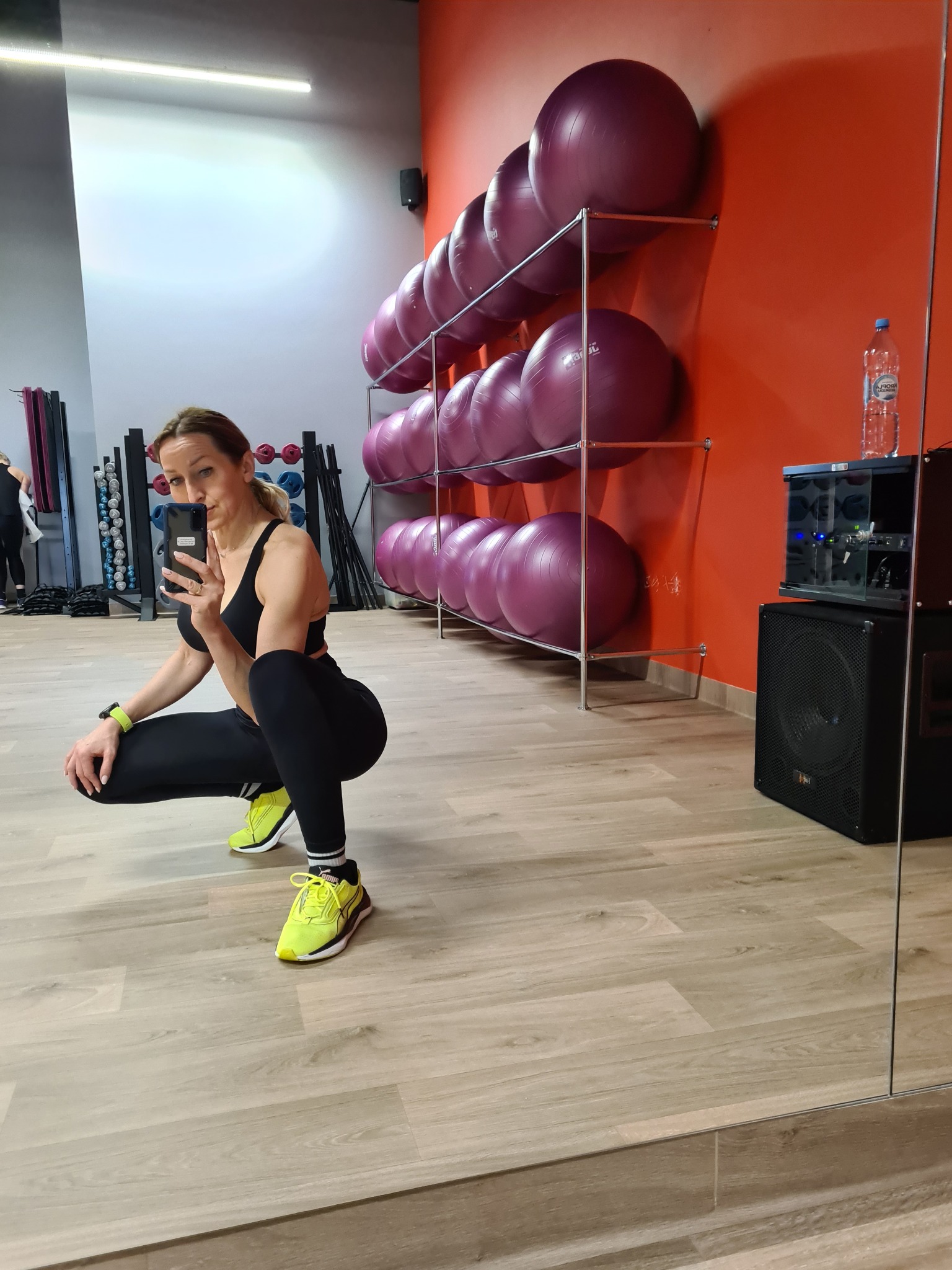 Kobieta w sportowym stroju robi selfie w siłowni na tle ściany z piłkami do ćwiczeń i lustra. Uwagę przykuwają neonowe buty i zegarek sportowy.