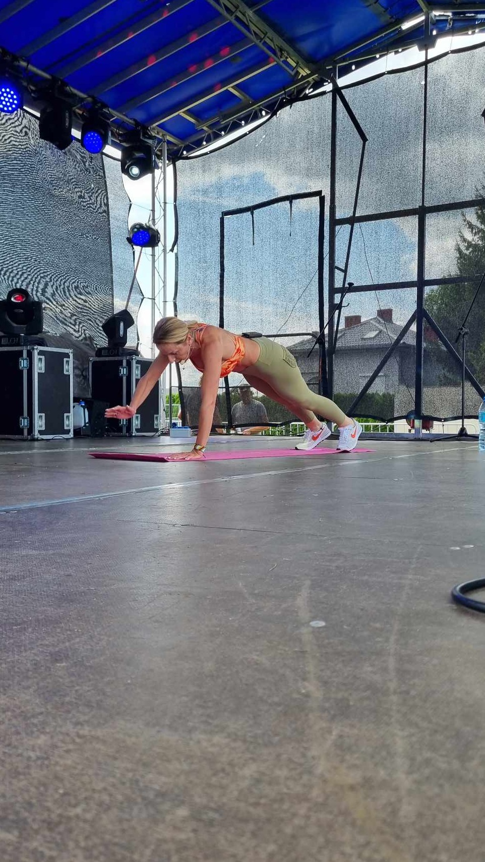 Trenerka personalna w pomarańczowym topie i legginsach wykonuje ćwiczenie na macie na scenie, unosząc jedną rękę. W tle sprzęt sceniczny i budynek.