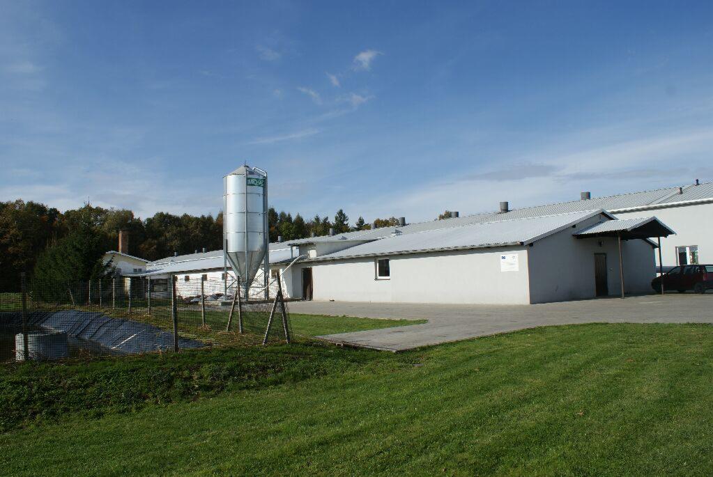 Widok zewnętrzny fermy drobiu z silosem paszowym, budynkami gospodarczymi i zielonym trawnikiem na pierwszym planie.