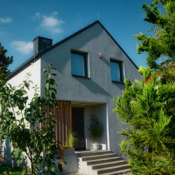 Verte Pracownia Projektowa - Nowoczesny dom jednorodzinny z szarą elewacją, dwuspadowym dachem i minimalistycznym wejściem, otoczony zielenią drzew i krzewów w słoneczny dzień.