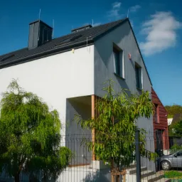 Nowoczesny dom jednorodzinny z dwuspadowym dachem w kolorze grafitowym, z jasnoszarą elewacją i elementami w kolorze białym, widoczny od strony ogrodu za metalowym ogrodzeniem.