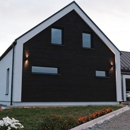 Verte Pracownia Projektowa - Nowoczesny dom jednorodzinny z czarną drewnianą elewacją i białymi ścianami, oświetlony lampami elewacyjnymi, z widocznym ogrodem i podjazdem z szarego żwiru.