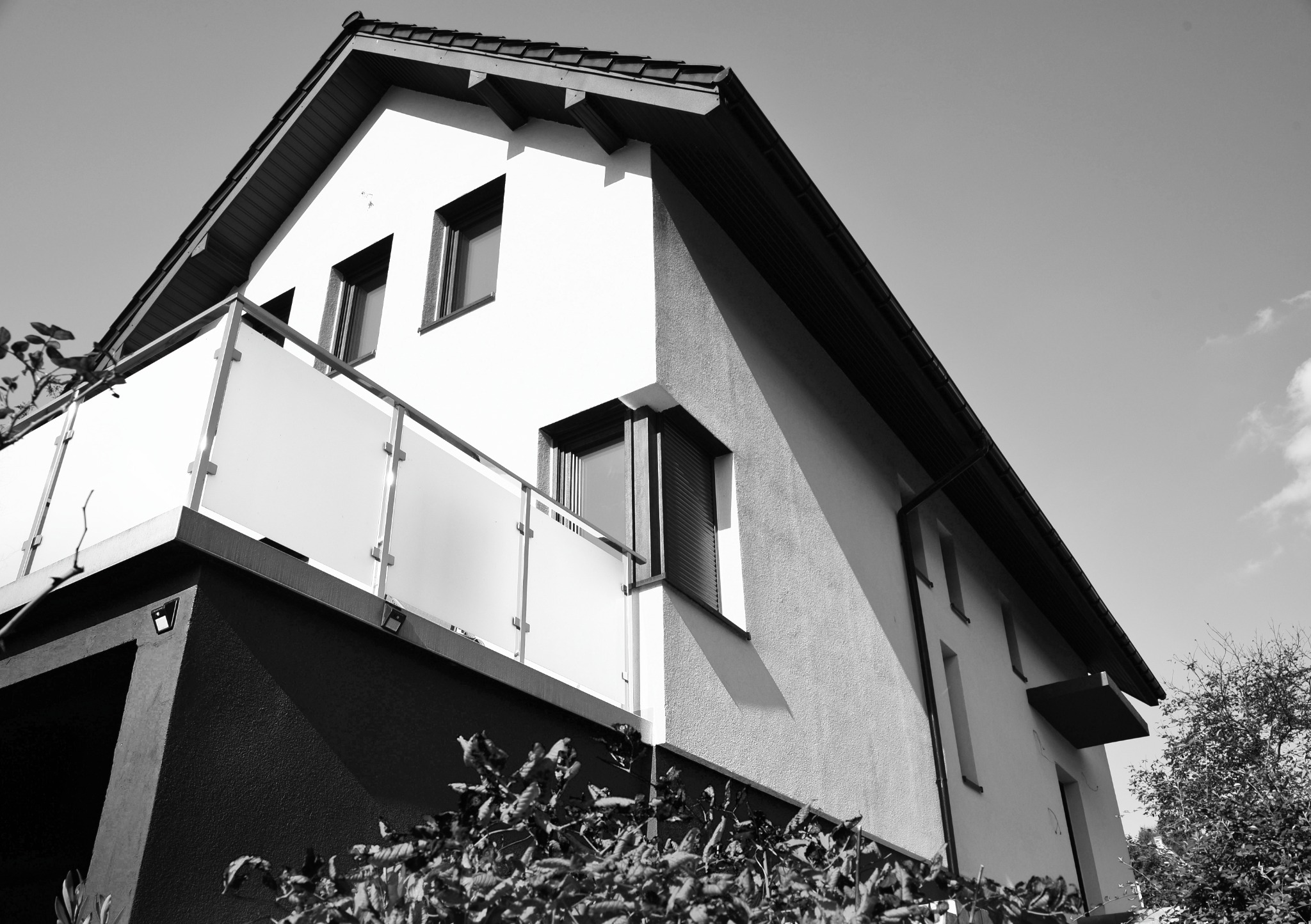 Czarno-białe zdjęcie domu z balkonem i nowoczesnymi oknami, widok od dołu, podkreślający geometrię budynku i kontrast między jasną elewacją a ciemnym dachem.