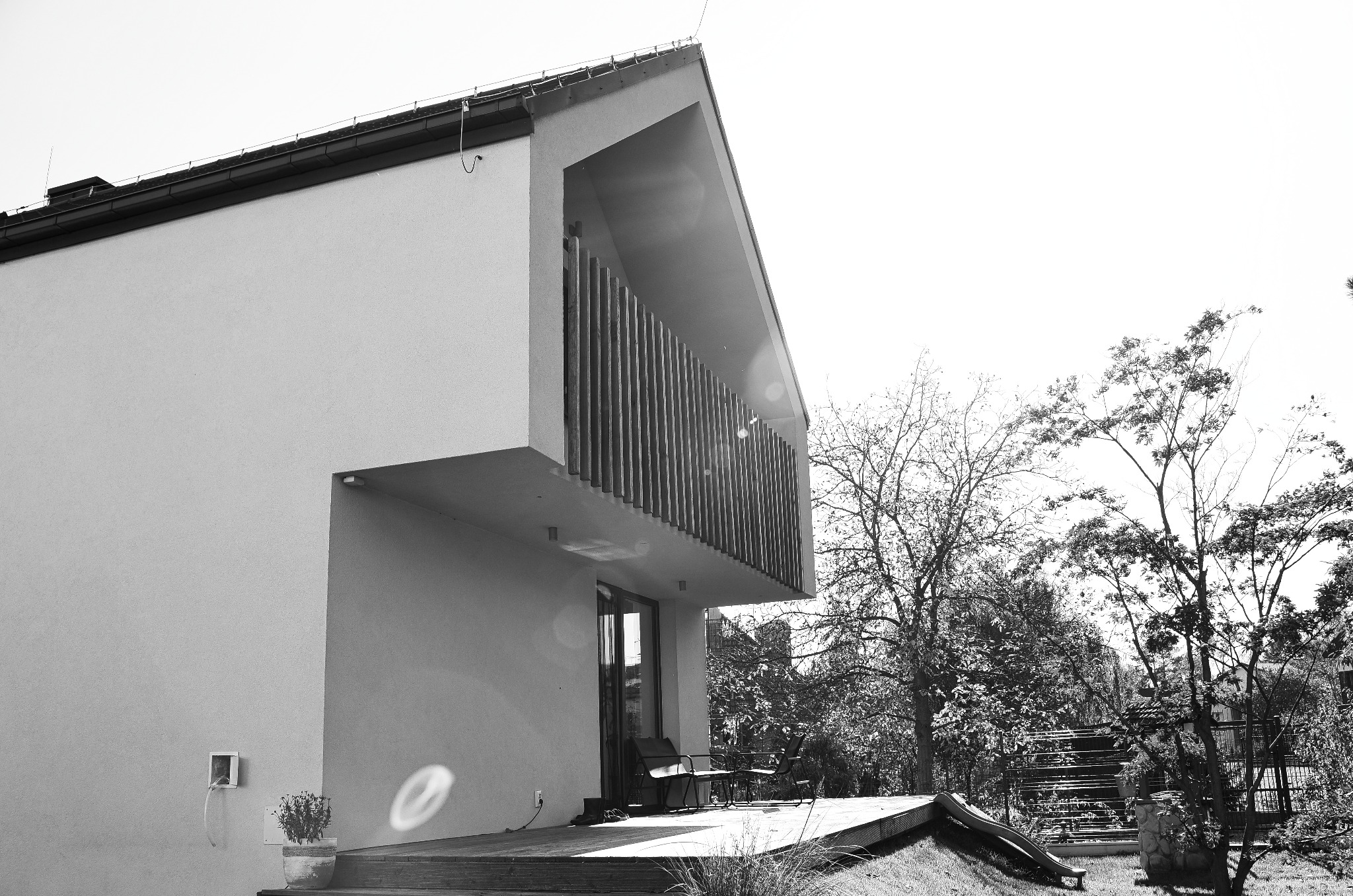 Czarno-białe zdjęcie nowoczesnego domu z balkonem z drewnianymi pionowymi panelami, widok z zewnątrz na taras z meblami ogrodowymi i zielenią w tle.