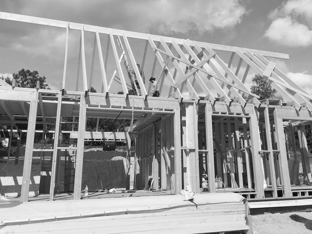 Czarno-białe zdjęcie w trakcie budowy domu szkieletowego, widoczne drewniane elementy konstrukcyjne, w tle drzewa i pochmurne niebo, robotnicy na drabinie montujący dach.