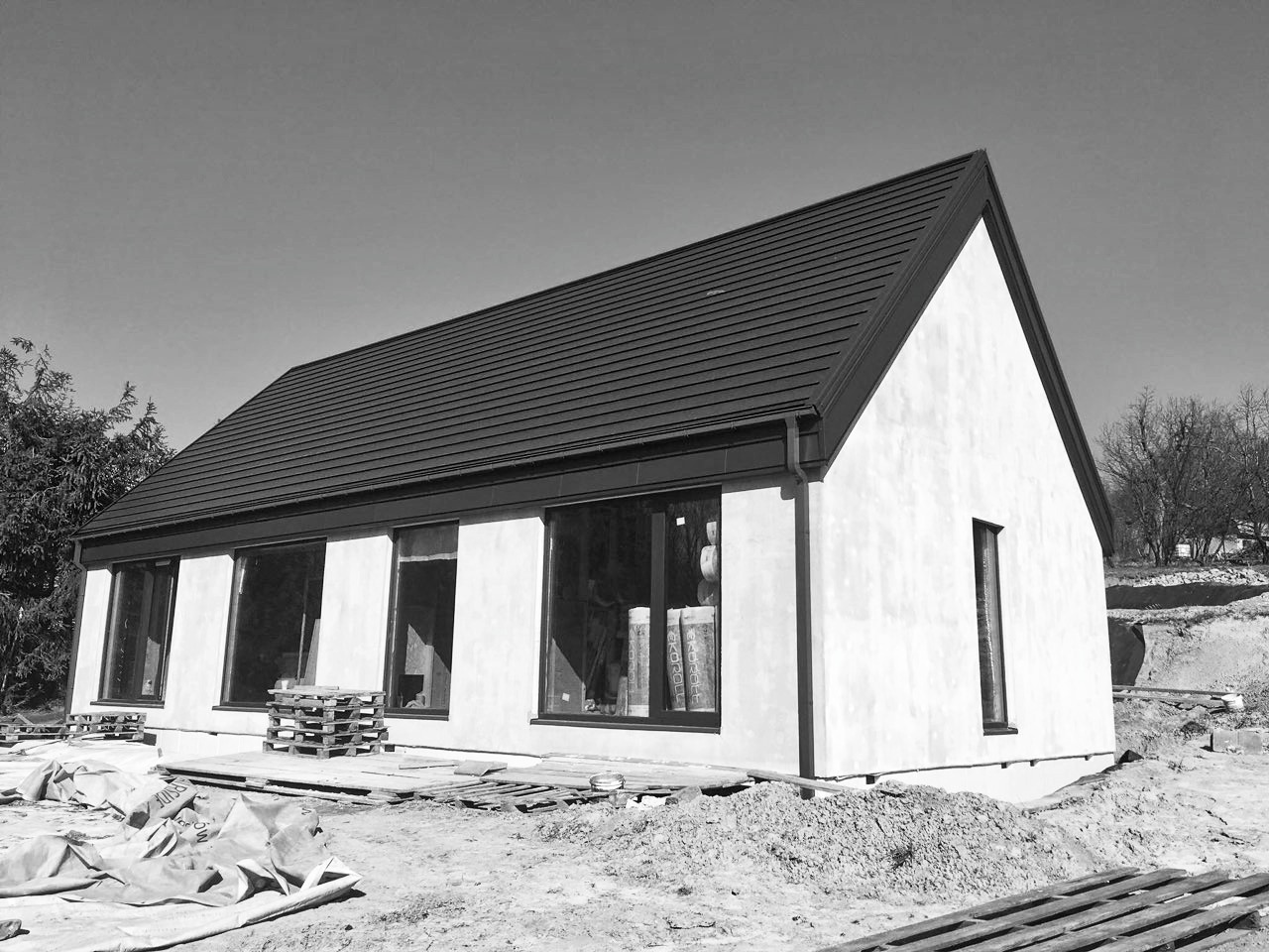 Czarno-białe zdjęcie domu w trakcie budowy, z widocznymi paletami, materiałami budowlanymi i ziemią wokół, okna częściowo zasłonięte folią.