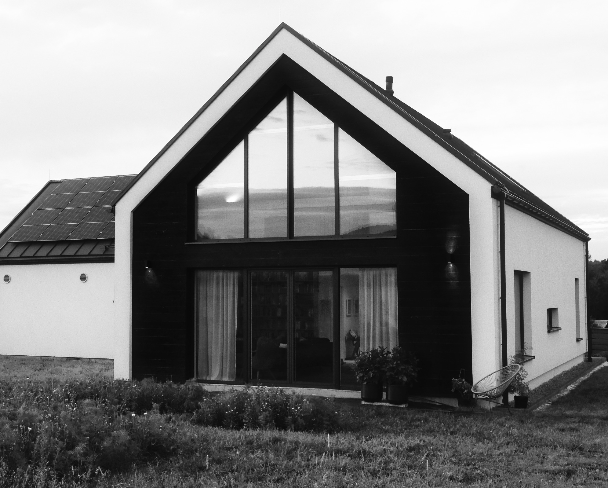 Czarno-białe zdjęcie nowoczesnego domu z dużymi oknami, panele słoneczne na dachu, otoczenie zarośnięte trawą i kwiatami.