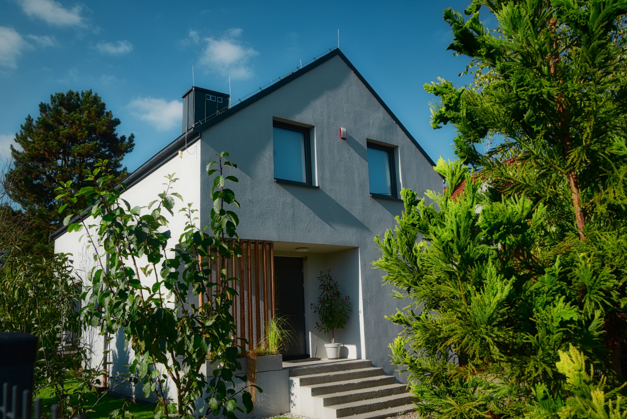 Nowoczesny dom jednorodzinny z szarą elewacją, dwuspadowym dachem i minimalistycznym wejściem, otoczony zielenią drzew i krzewów w słoneczny dzień.