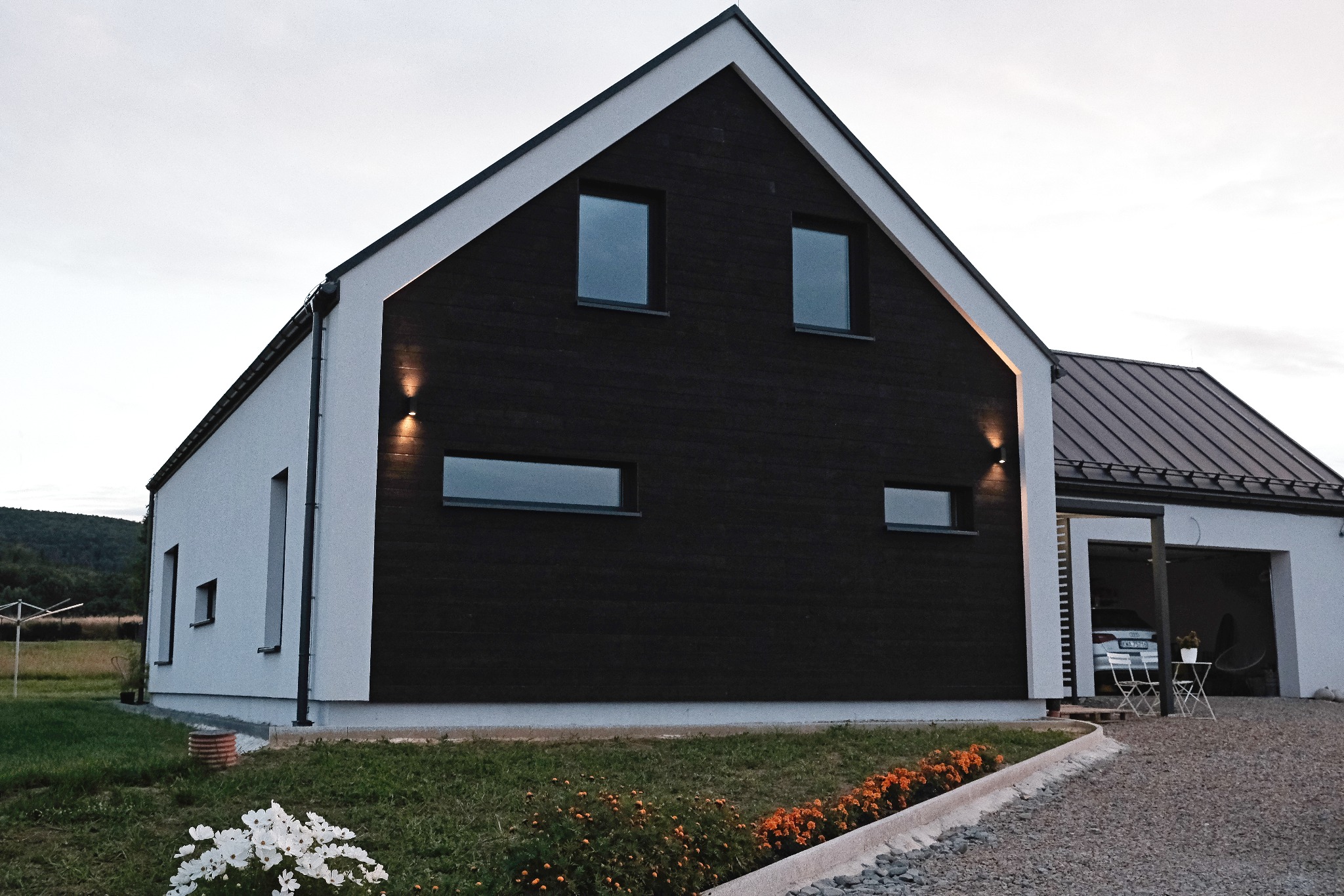 Nowoczesny dom jednorodzinny z czarną drewnianą elewacją i białymi ścianami, oświetlony lampami elewacyjnymi, z widocznym ogrodem i podjazdem z szarego żwiru.