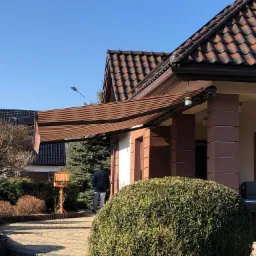 Brązowa, rozłożona osłona przeciwsłoneczna nad wejściem do domu z ciemnobrązową dachówką, na tle błękitnego nieba i osoby idącej w kierunku domu.