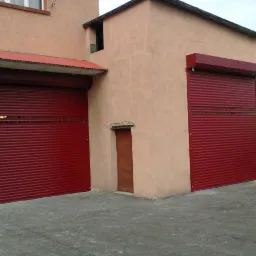 Dwie czerwone bramy garażowe rolowane w budynku o beżowej elewacji. Jedna brama zadaszona, druga bez zadaszenia. Małe drzwi wejściowe obok większej bramy.